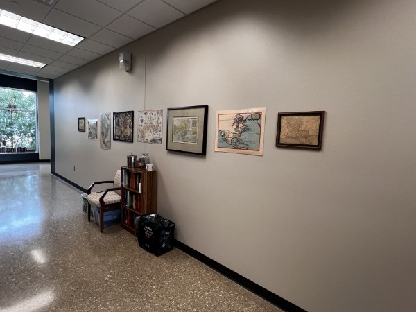 A row of framed and unframed maps along a hallway wall above a chair and small bookshelf.