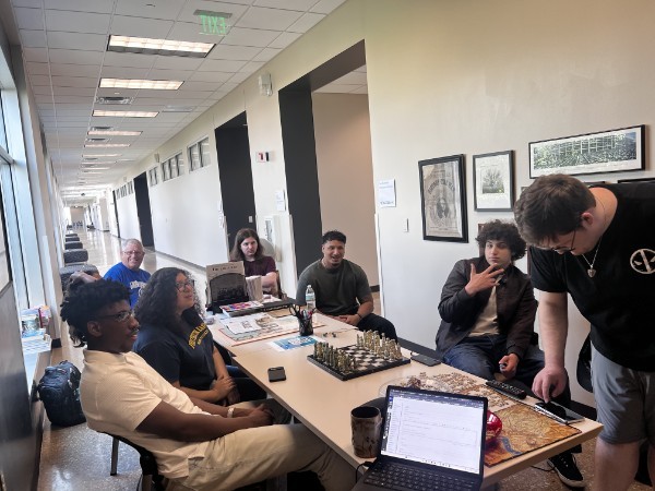 A group gathered around tables with a chessboard, books, a laptop, and a map.